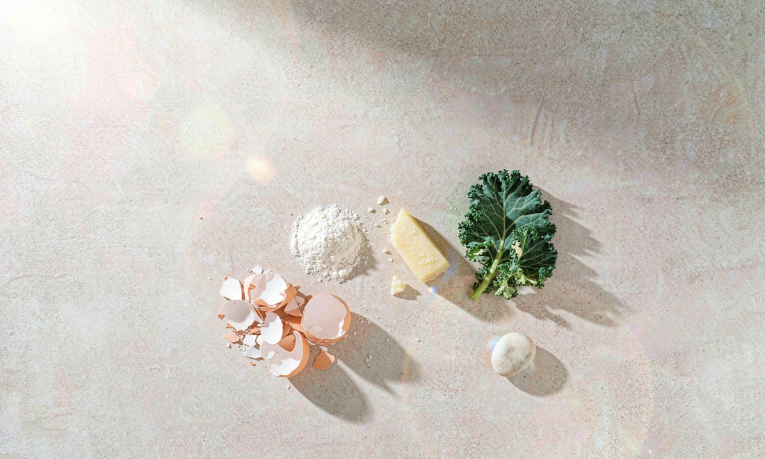 Flat lay of eggshells, mineral powder, cheese, kale and a pebble arranged on stone in warm sunlight