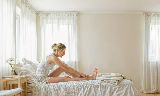 Woman gently stretching her calf on the edge of a pale linen bed at dusk with a glass of water on the nightstand