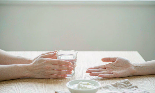 Two relaxed hands on a light oak table beside a glass of water and a small bowl of yogurt