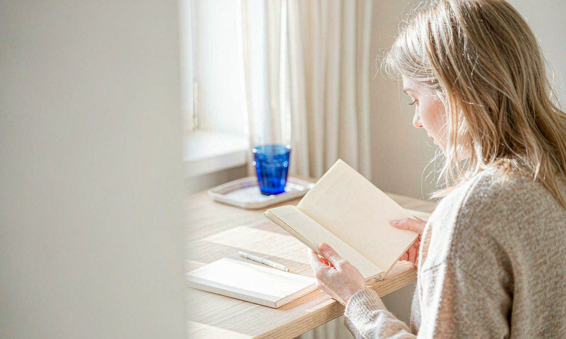 Woman with blonde hair seen from behind reading a notebook at a sunlit desk with a cobalt blue glass