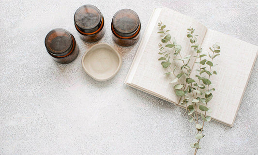 Overhead flat lay of plain amber jars, a ceramic dish, an open linen notebook and eucalyptus on a pale stone counter
