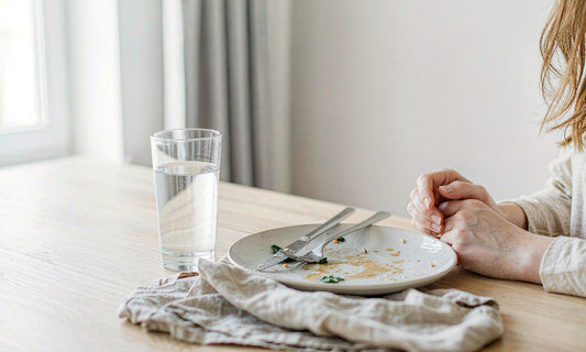 Relaxed hands resting beside a finished lunch plate and a glass of water on a pale oak table in soft daylight