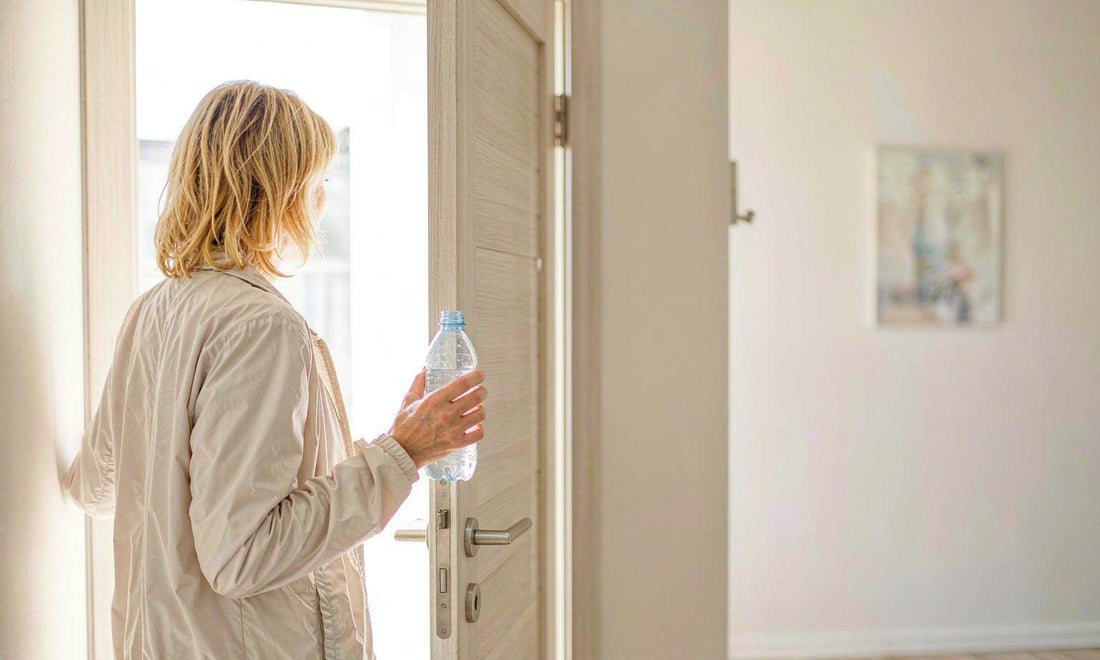 Woman with blonde hair seen from behind stepping out a sunlit door holding a water bottle