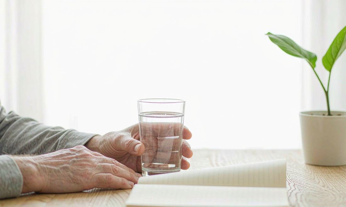 Fair-skinned mature man's hands holding a glass of water at a sunlit wooden desk