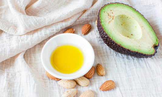 Golden oil droplets in a white ceramic dish with almonds and avocado half on pale linen, overhead view