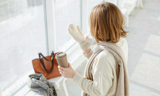 Person from behind pulling on warm gloves by a frosted window with a travel cup