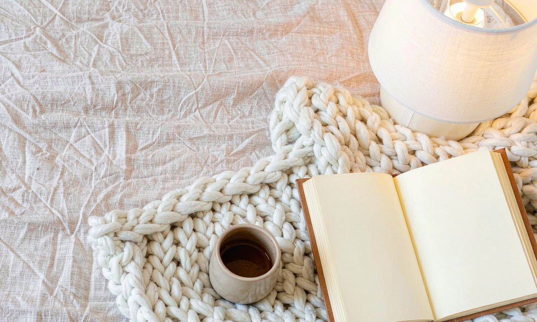 Overhead flat lay of a pale linen bed corner with a knit blanket, ceramic cup and open journal in warm lamp light