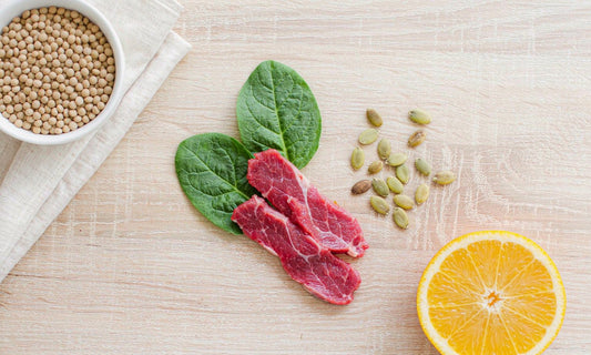 Overhead flat lay of iron-rich foods with lentils spinach beef pumpkin seeds and a halved orange on pale oak