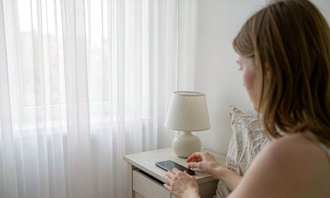 Person setting a phone face down on a nightstand beside a lamp in a bright bedroom