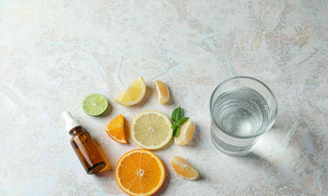 Overhead flat-lay of a glass dropper bottle, fresh citrus slices and water on pale marble