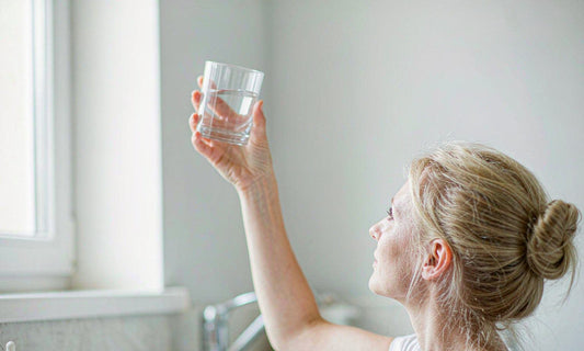 Woman in a bright kitchen raising a glass of water toward soft morning window light