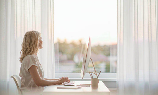 Woman with blonde hair taking a screen break by a bright window at a home desk in soft morning light