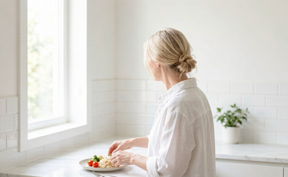 A woman in her fifties preparing a vegetable and grain plate at a bright kitchen counter, seen from behind