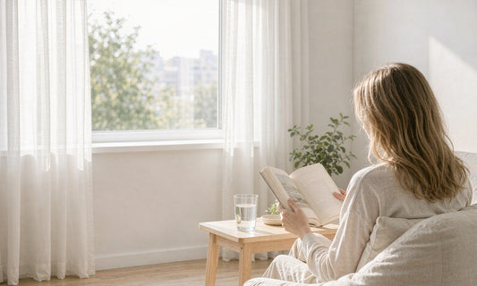 Woman seen from behind reading by a bright sunlit window with a glass of water nearby