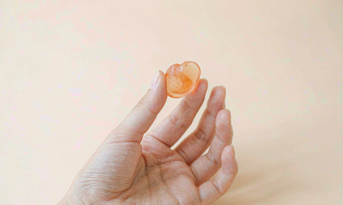 A fair skin hand holding one translucent orange gummy between two fingers against a soft cream background