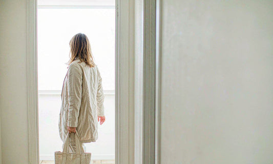 A woman seen from behind stepping through a bright home doorway with a canvas tote, morning light filling a pale hallway