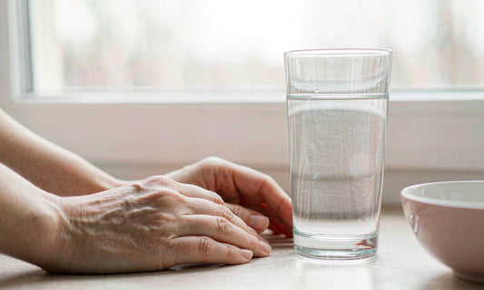 A pair of relaxed hands resting beside a tall glass of water and a small ceramic bowl on a pale kitchen counter in soft window light