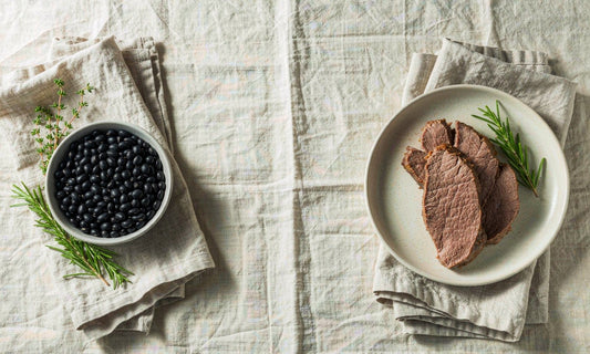 Overhead comparison of a bowl of black beans and a plate of sliced lean beef on a linen tablecloth