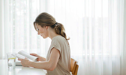 Woman with light brown hair reading at a bright desk beside a glass of water in soft daylight