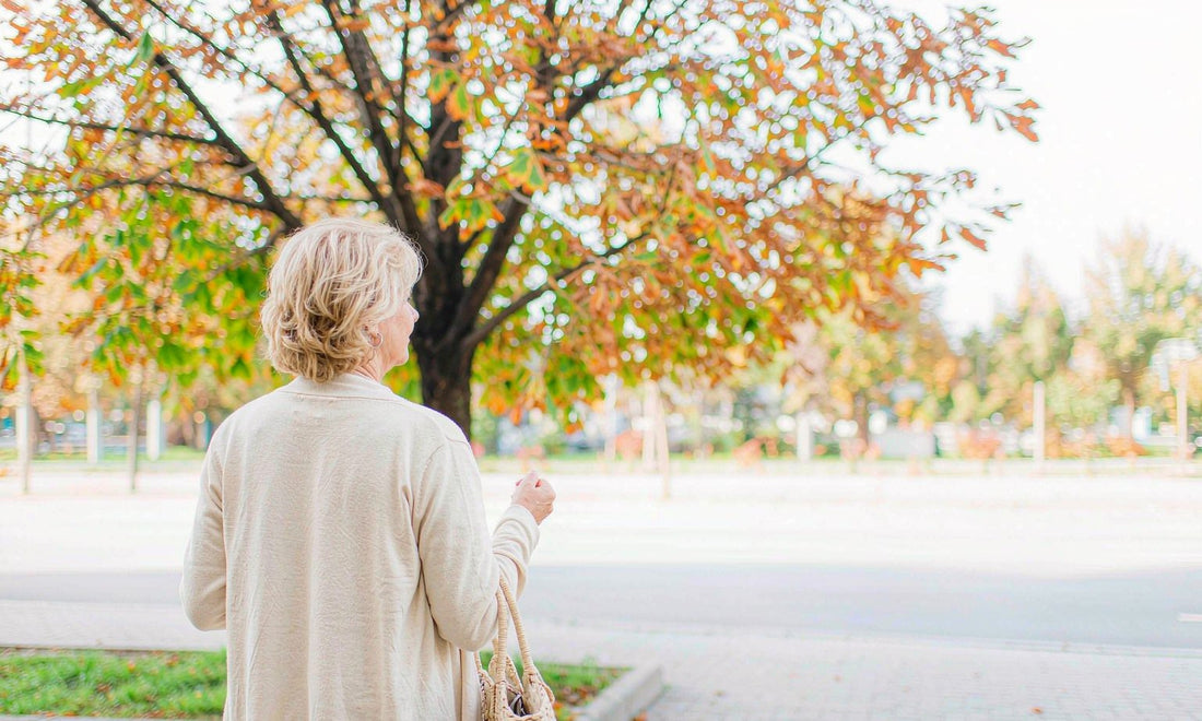 Woman in her fifties with blonde hair and a cream cardigan walking a sunlit autumn sidewalk, seen from behind