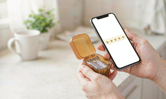Two hands with fair light skin holding an amber supplement tray next to a phone showing a five-star review grid, representing customer feedback in a bright kitchen
