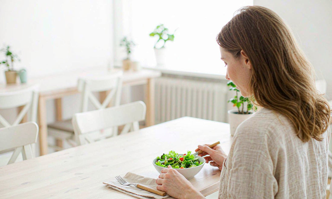 Woman with light brown hair seen from behind eating a salad slowly at a bright cafe table