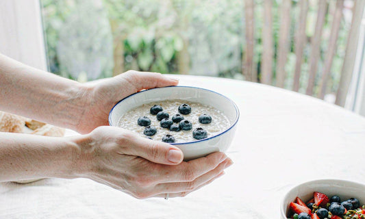 A pair of hands holding a ceramic bowl of creamy oatmeal topped with blueberries on a bright table