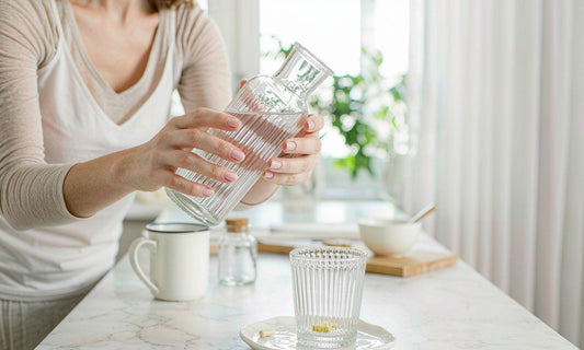 Hands pouring water beside a saucer of capsules in a bright morning kitchen, a simple daily routine