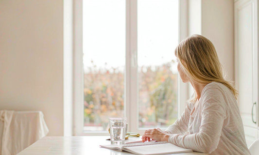 Woman seen from behind planning a calm fall morning routine at a sunlit kitchen table with water