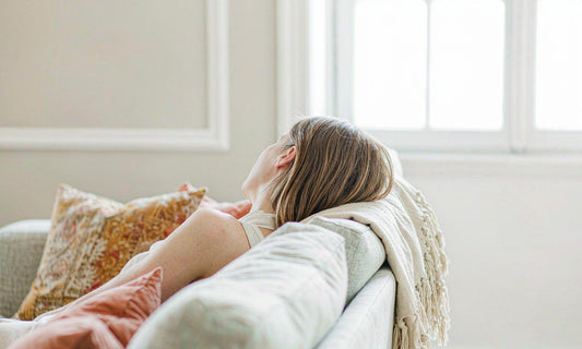 A woman resting quietly on a pale sofa beside a bright window in soft afternoon light