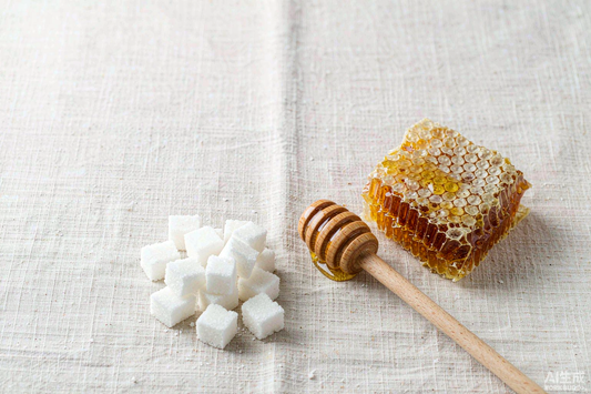Honeycomb with a wooden honey dipper beside white sugar cubes on pale linen, comparing two sweeteners