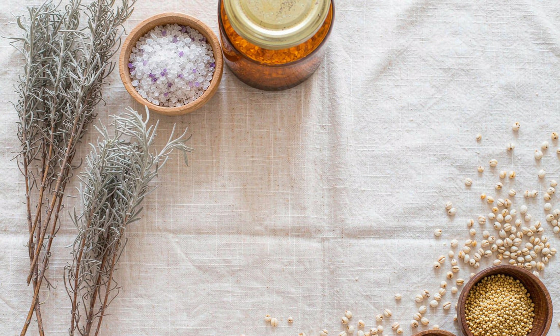 Overhead arrangement of dried herbs, coarse sea salt, amber jars and toasted grain clusters on pale linen, evoking whole-food nutrient density