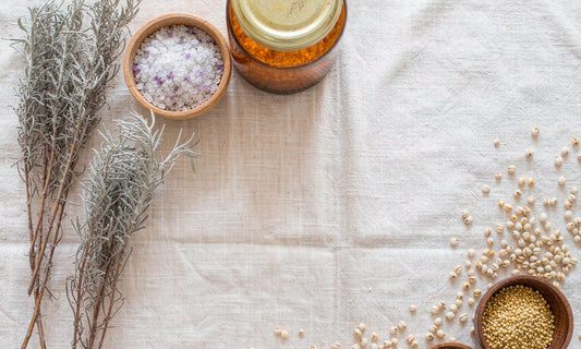 Overhead arrangement of dried herbs, coarse sea salt, amber jars and toasted grain clusters on pale linen, evoking whole-food nutrient density