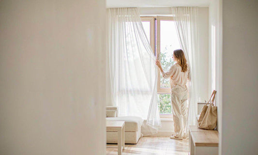 A woman seen from behind at a sunlit sheer-curtained window in a bright minimal living room