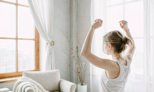 Woman with light brown hair stretching by a bright window in a calm morning room, seen from behind