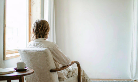 Person seen from behind relaxing in a linen armchair by a sunlit window with a mug and book on a side table