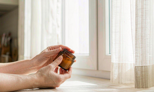 Fair skinned hands holding an amber supplement jar by a bright kitchen window with sheer curtains