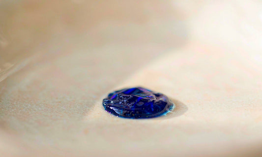 Macro close-up of a single deep blue liquid droplet on a pale ceramic surface with soft bokeh