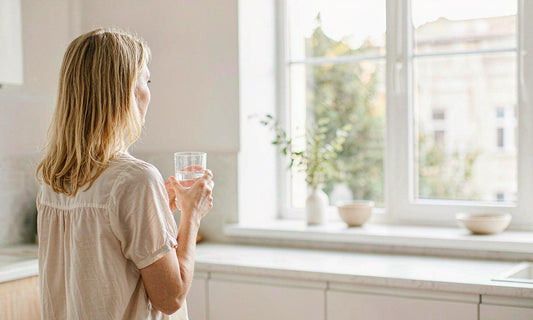Woman seen from behind holding a glass of water at a bright kitchen window in soft morning light
