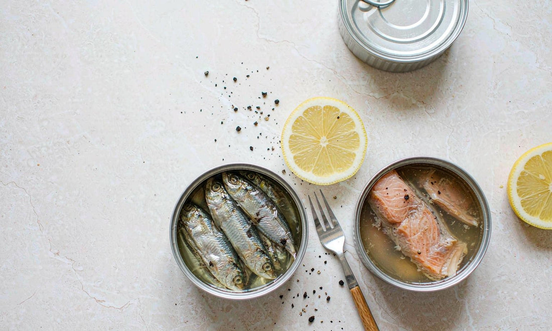 Open tins of sardines and salmon with lemon slices and black pepper on a pale stone counter