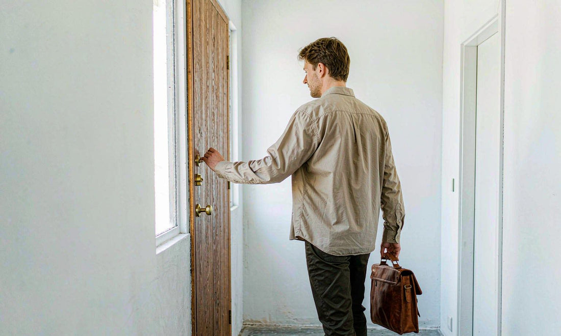 A Western man seen from behind holding a leather work bag in a bright pale home hallway, morning light through a side window