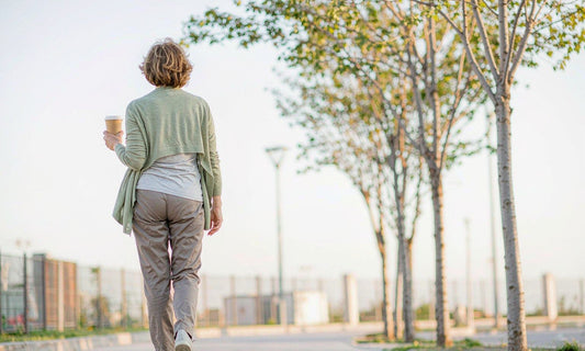 Woman seen from behind walking on a sunny sidewalk in a layered cardigan holding a coffee cup