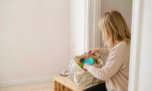 A blonde woman seen from behind packing a lunch container into a tote in an entryway
