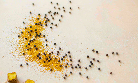 Golden turmeric powder and scattered black peppercorns arranged on a pale cream ceramic surface with dried root slices