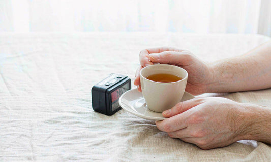 Fair-skinned hands holding a warm cup of tea beside a small alarm clock on pale linen bedding