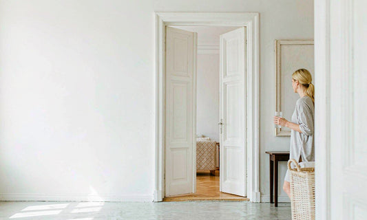 Woman with blonde hair seen from behind walking through a bright hallway holding a glass of water