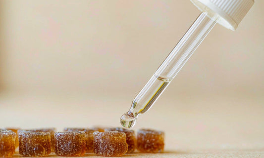 Macro shot of a dropper releasing a clear liquid drop next to a row of amber gummies on a cream surface