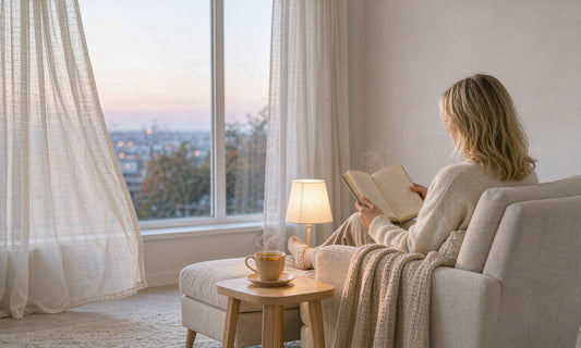 A woman seen from behind reading in a cozy armchair beside a warm lamp and herbal tea at dusk