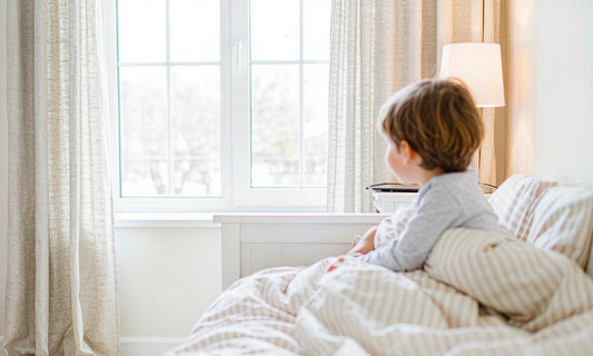 Child seen from behind in a cosy bedroom at dusk with a tablet resting face-down beside a warm lamp