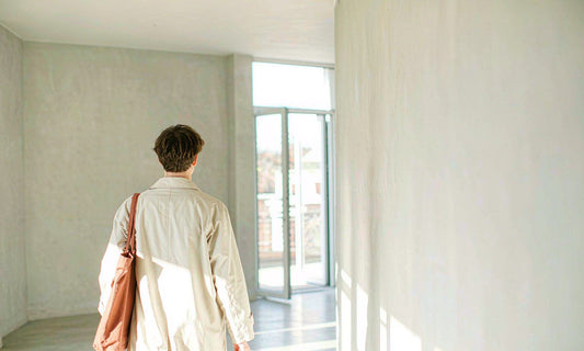 Person seen from behind in an ivory coat with a canvas tote walking through a bright minimal hallway toward glass doors